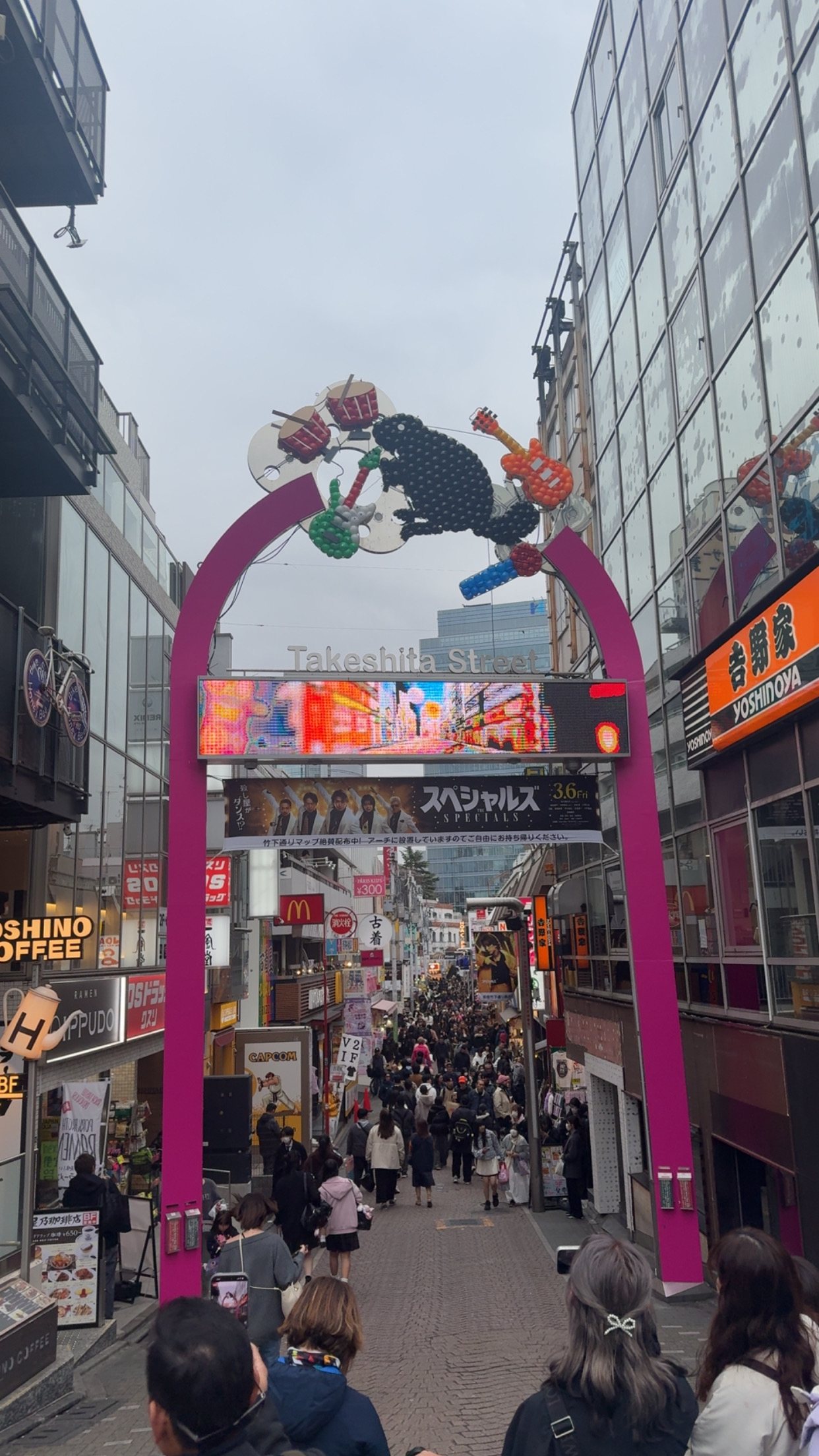 Looking straight down the Takeshita Street corridor from inside the crowd at eye height: the pink arch overhead, shoppers