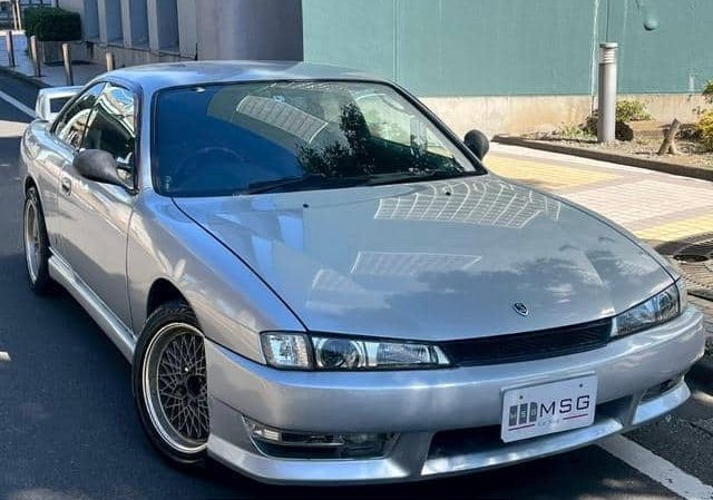 Tight crop on the front three-quarter of the silver S14 Silvia sitting on a Japanese street with its dealer plate visible